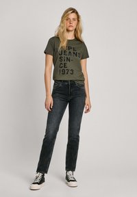 Zelena majica s crnim natpisom "PEPE JEANS SINCE 1973," u kombinaciji s tamnim trapericama i crnim visokim tenisicama s bijelim potplatima.