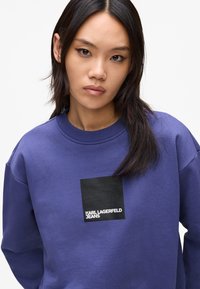 Purple sweatshirt with a ribbed neckline and long sleeves. Features a black square patch with "KARL LAGERFELD JEANS" printed in white.