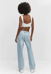 Top de punto blanco corto con bordes ondulados, combinado con jeans de pierna ancha de cintura alta en azul claro. Zapatos planos blancos. Fondo liso.