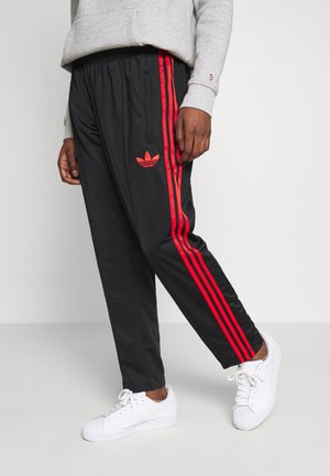 Personne portant un pantalon de jogging noir avec des bandes rouges et le logo Adidas, un sweat gris et des baskets blanches, debout sur un sol clair devant un mur blanc.