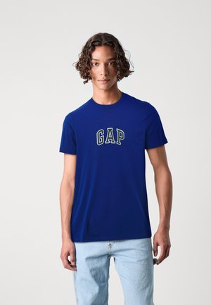 Tume sinine puuvillane T-särk, ümarate kaelustega, millel on rohelisest ja valgest brodée "GAP" logo, kantud heledate siniste teksadega.