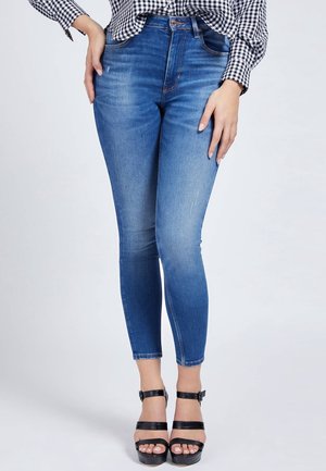 Kobieta ubrana w niebieskie, dopasowane jeansy z wysokim stanem, czarne sandały na obcasie z paskami oraz czarno-białą, kratowaną koszulę z długim rękawem.