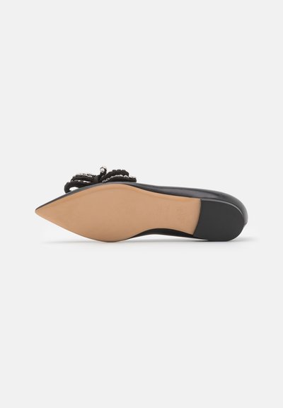 Zapato plano negro de punta con parte superior de tejido texturizado, lazo decorativo y sutiles acentos metálicos. Suela de goma marrón claro.