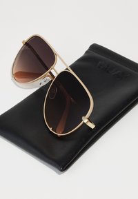 Gafas de sol aviador doradas con lentes oscuros degradados, reposando sobre un estuche negro texturizado con "QUAY" grabado.