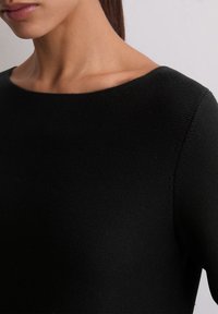 Femeie îmbrăcată cu un top negru texturat, cu gât barcaj, arătată din umeri până la bărbie, pe un fundal deschis simplu.