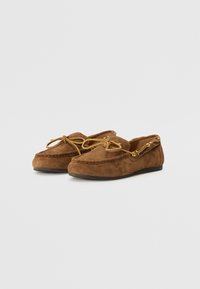 Scarpe mocassino in suede marrone, con punta arrotondata, lacci frontali e accenti dorati. Suola piatta e texture morbida.