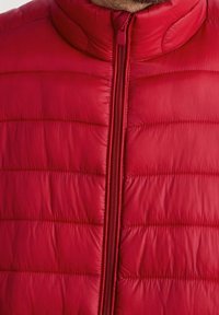 Červená prošívaná vesta s vysokým límcem a celopropínacím zipem vpředu. Vyznačuje se prošívanou texturou s horizontálními švy a lesklým, hladkým povrchem.