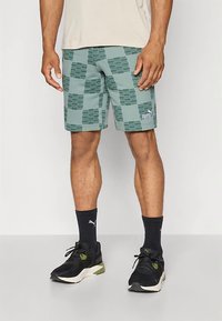 Πράσινα σορτς με σχέδιο καρό και αντίθετο λογότυπο, κατασκευασμένα από ελαφρύ υλικό, φορεμένα με μαύρα αθλητικά παπούτσια και κάλτσες.