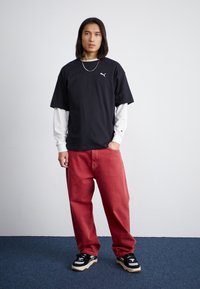 Tricou negru cu mânecă scurtă, suprapus peste un tricou alb cu mânecă lungă, asortat cu pantaloni roșii largi și sneakers negri cu accente albe.