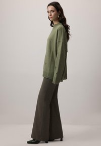 Olivegreenowy, oversize sweter z prążkowanym dołem i przetartymi detalami, zestawiony z brązowymi spodniami o wysokim stanie w fasonie dzwonów oraz czarnymi szpilkami.