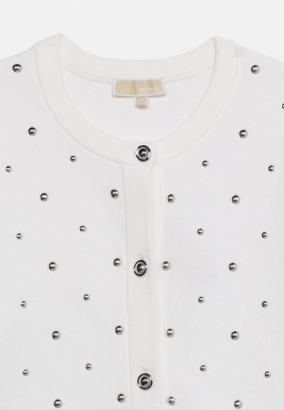 Λευκή πλεκτή ζακέτα με στρογγυλή λαιμόκοψη, ασημένια κουμπιά κατά μήκος του μπροστινού μέρους και ομοιόμορφα τοποθετημένα μικρά ασημένια καρφιά ως διακόσμηση.