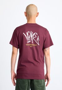 Pessoa careca com brincos, vestindo uma t-shirt Nike cor bordeaux com grandes letras brancas em estilo graffiti e um pequeno logótipo amarelo nas costas, e calças verdes.