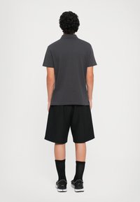 Mann med krøllete mørkt hår står med ryggen til, iført en mørkegrå polo-skjorte, svarte shorts, svarte sokker og svarte joggesko med hvite såler.