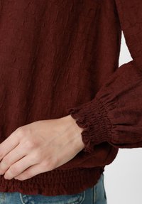 Χέρι που ακουμπά κοντά στη μέση, φοράει μπορντό υφασμάτινο μπλουζάκι με ελαστικά μανίκια και μπλε τζιν παντελόνι.