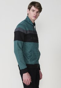 Sudadera de rayas en tonos verde azulado con un cuello alto, que presenta acentos horizontales en negro y gris, y un bolsillo frontal. La tela es texturizada y suave.
