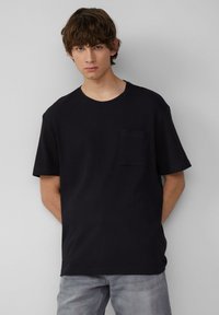 Czarny t-shirt z krótkim rękawem o prążkowanej fakturze, okrągłym dekolcie i kieszeni na piersi. Model stoi w jasnoszarych dżinsach na szarym tle.