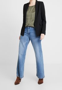 Blazer nero sopra un top verde oliva, abbinato a jeans a gamba larga blu chiaro e stivaletti con tacco marroni. Presenta un colletto tradizionale e tasche.