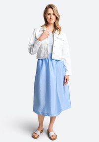 Weiße, kurze Jeansjacke über einem blauen, ärmellosen, knielangen Kleid. Das Kleid hat eine glatte Textur; das Model trägt beige Sandalen.
