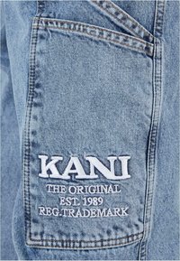 Tela de mezclilla azul claro con costuras visibles. Presenta un logo bordado en blanco "KANI" y el texto "EL ORIGINAL EST. 1989 MARCA REGISTRADA."