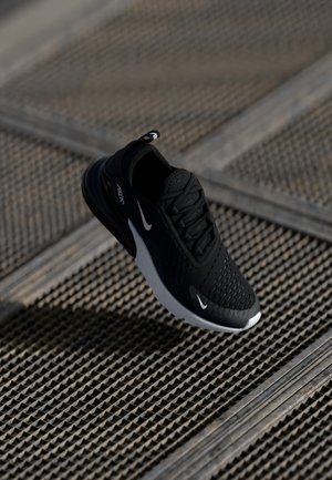 Baskets Nike Air Max 270 noires avec détails swoosh blancs reposant sur une surface en grille métallique texturée.