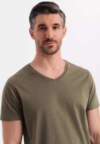 Olivově zelené tričko s výstřihem do V vyrobené z jemného materiálu, s krátkými rukávy a hladkým povrchem, které nosí muž s krátkými vlasy.