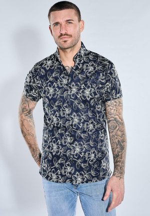 Man with short hair and tattoos on both arms wears a dark blue short-sleeve shirt with white floral patterns and light blue jeans.
