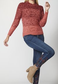Knitted sweater with a gradient of red hues, featuring a textured pattern. Paired with blue jeans showcasing colorful floral embroidery at the hem.