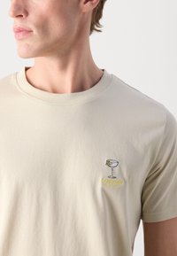 Beige pamut póló kerek nyakkal, rövid ujjú, és egy kis hímzett koktélpohárral, amelyen a "Lidberg's Cocktails" felirat áll.