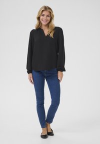Blouse noire à manches longues avec un col en V, associée à un jean ajusté bleu et des chaussures plates noires. Design simple, texture lisse, style décontracté.