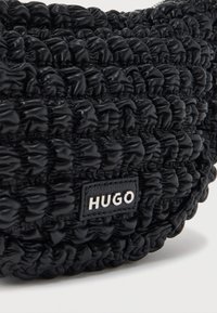 Κοντινό πλάνο μιας μαύρης υφασμάτινης τσάντας με μικρή ορθογώνια μαύρη δερμάτινη ετικέτα σφραγισμένη με λευκά γράμματα «HUGO».