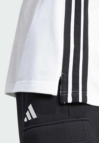Bílý sportovní tričko s černými pruhy po stranách, se bočním rozparkem. Černé kalhoty s bílým logem Adidas na stehně.