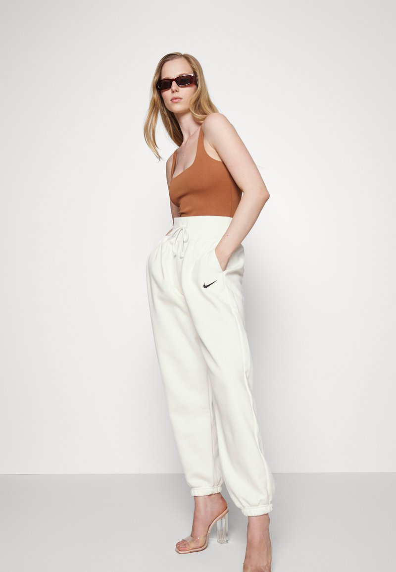 Top de tirantes finos marrón ajustado, combinado con pantalones de chándal de forro polar blancos con cintura elástica y logo negro de Nike. Sandalias de tacón transparente.