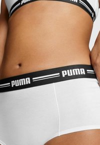Nahaufnahme des Mittelbereichs einer Person, die weiße Puma-Unterwäsche mit einem schwarzen Bund trägt, auf dem sich das Puma-Logo und Streifen befinden.