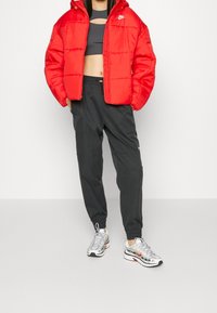 Röd puffjacka med huva, svart kort topp, grå sweatpants och silverfärgade sneakers med röda accenter. Slät textur och avslappnad passform.