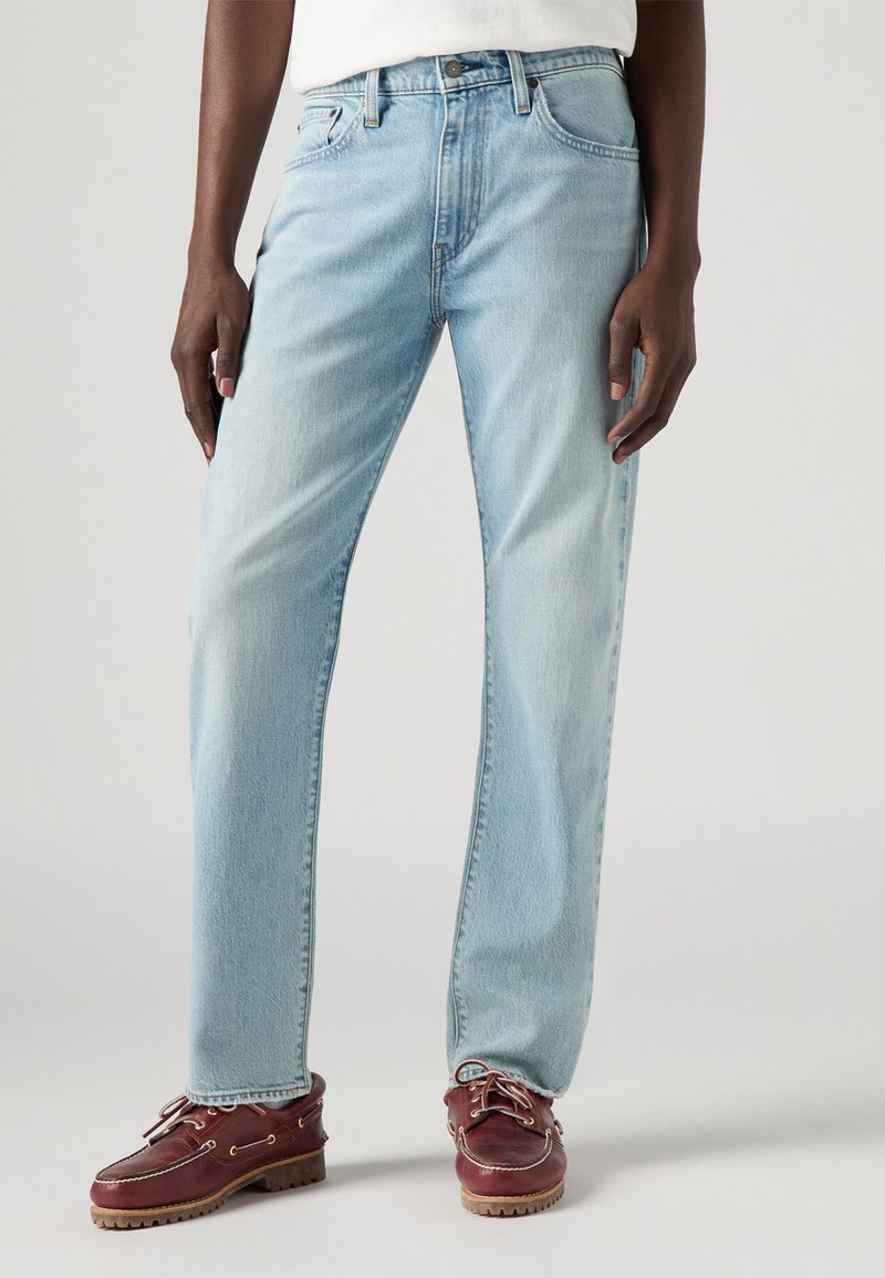 Jeans en denim bleu clair avec une coupe droite, dotés de poches avant et arrière et de détails de couture subtils, associés à des chaussures bateau rouge foncé.