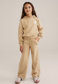 Beige huppari, jossa on valkoinen kukkakuvio, yhdistettynä matching beige-housuihin, joissa on sivusaumat ja tekstityksillä. Vaaleanpunaiset lenkkarit viimeistelevät asun.
