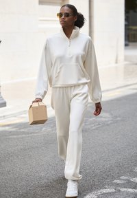 Biely, textúrovaný pulóver na zips a zodpovedajúce nohavice na jogging. Model drží malú hnedú krabičku, má na sebe slnečné okuliare a biele tenisky.