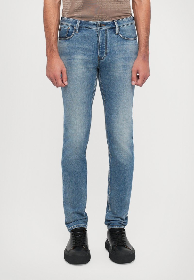 Világoskék farmernadrágok slim fit fazonnal rendelkeznek, finom kopással. Öt zsebbel és hagyományos gombos záródással készültek. Fekete sportcipővel viselik.