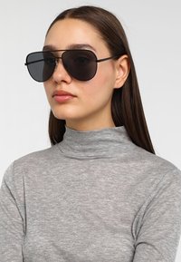 Ochelari de soare aviator negri cu lentile fumurii, rame subțiri din metal, purtați cu un top cu gât înalt, de culoare gri deschis, cu mâneci lungi, textură netedă.