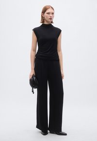 Top preto sem mangas com gola alta, detalhes franzidos na cintura, combinado com calças largas pretas e uma pequena mala preta.