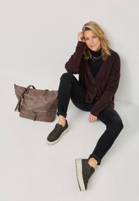 Dunkelrote Strickjacke über einem schwarzen Rollkragenpullover, schwarze Jeans und strukturierte olivgrüne Sneaker mit Leopardenmuster. Braune Tragetasche mit Reißverschluss und Quaste.