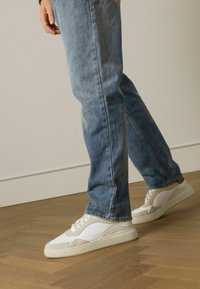Witte en beige sneakers met gestructureerde panelen, vetersluiting en rubberen zolen, gecombineerd met lichtblauwe denim jeans tegen een houten vloer.