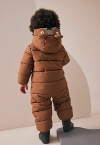 Brązowy kombinezon puchowy o pikowanej fakturze, z kapturem z motywem misia i uszami, w połączeniu z czarnymi gumowymi butami. Widok od tyłu.