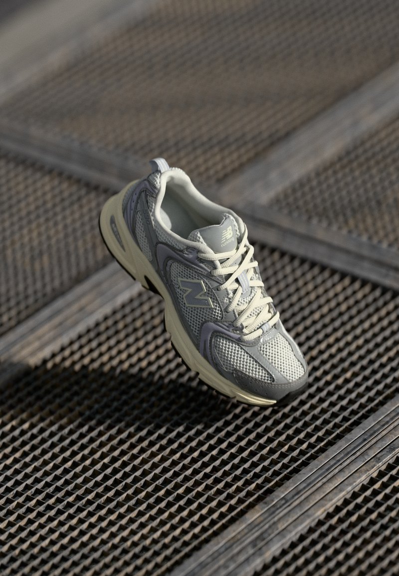 Chaussure de sport en gris clair et crème, fabriquée en matériau en maille, avec un col rembourré, un design à lacets et une semelle texturée pour une meilleure adhérence.
