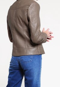 Veste en cuir marron avec finition texturée, coupe ajustée et accents cousus, portée sur un jean en denim bleu. Vue arrière visible.