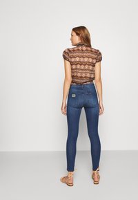 Mörkblå skinny jeans i kombination med en mönstrad kortärmad blus, med hög midja och bakfickor med en etikettlogotyp. Robust sandaler ingår.