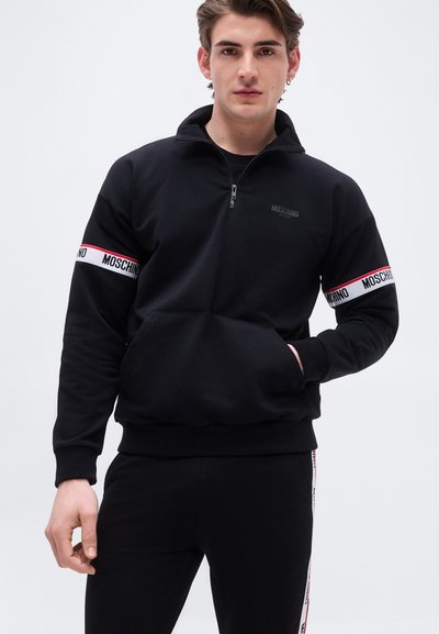 Νεαρός άνδρας φοράει μαύρο φούτερ Moschino με φερμουάρ στο μισό μήκος, μπροστινή τσέπη και λωρίδες με λογότυπα στα μανίκια, καθώς και αντίστοιχο μαύρο παντελόνι με λωρίδες λογότυπων στο πλάι.