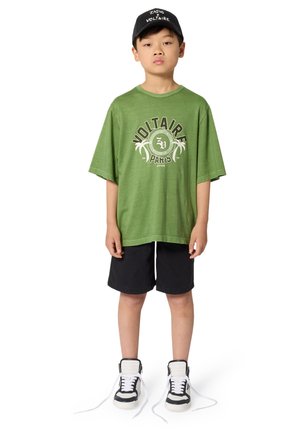Groen oversized t-shirt met een bedrukte "VOLTAIR"-print, zwarte korte broek en witte sneakers met zwarte accenten, gecombineerd met een zwarte pet.