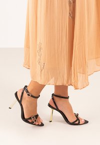 Sandalias de tacón alto de piel sintética negra con tiras finas, un tacón en tono dorado y detalles decorativos en hebilla. Combinadas con un vestido fluido color nude.