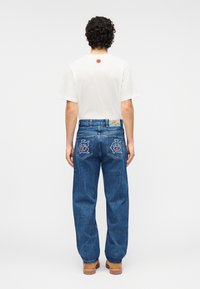 Μπλε τζιν παντελόνι με χαλαρή εφαρμογή, με κεντημένα σχέδια στις τσέπες και πολύχρωμο μπαλώμα στην πίσω μέση. Λευκό μπλουζάκι.